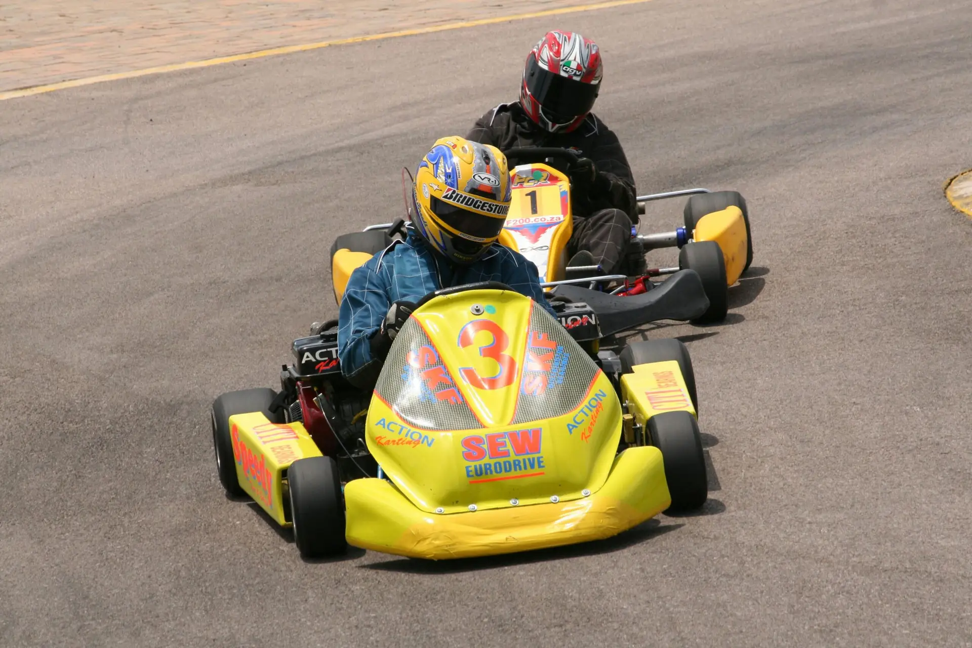 Two racers in vibrant go-karts compete on an outdoor track wearing safety gear.